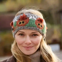 A woman wearing a knitted headband with crooked orange and brown bear faces, green panels, and gray trim, photographed outdoors with a blurred natural background.