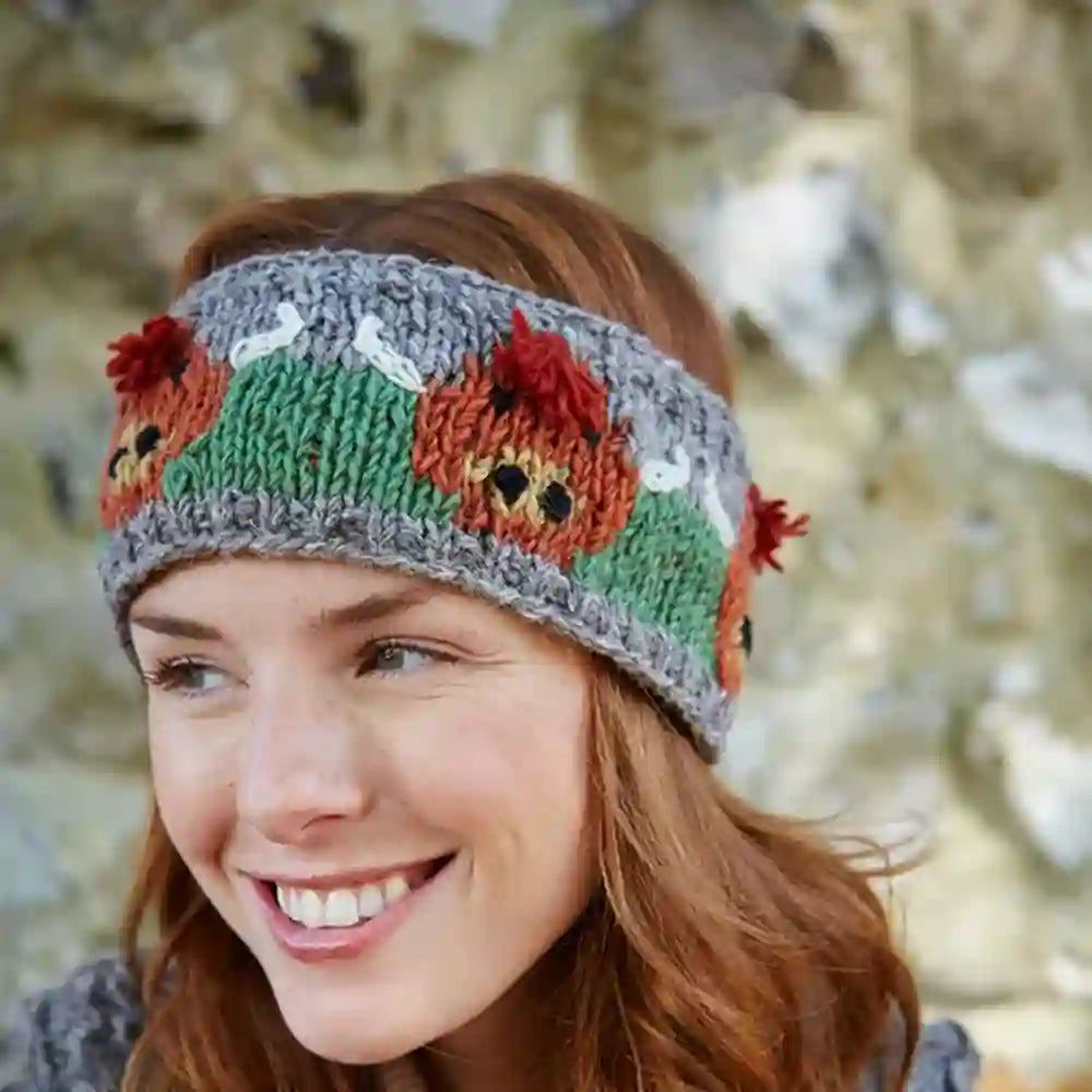 A smiling woman wearing a knitted headband with a pattern of green panels, red pom-poms, and orange-and-brown fox faces on a blurred natural stone background.