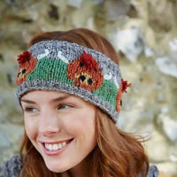 A smiling woman wearing a knitted headband with a pattern of green panels, red pom-poms, and orange-and-brown fox faces on a blurred natural stone background.
