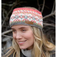 A woman wearing a knitted winter headband with a multicolor Fair Isle pattern of pink, orange, white, and gray, featuring a small brown leather brand patch on the front.