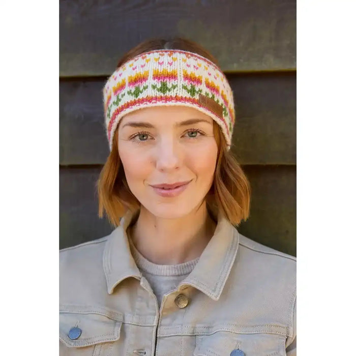 A woman wearing a knitted headband with a colorful floral pattern, paired with a light beige denim jacket, set against a dark wooden background.