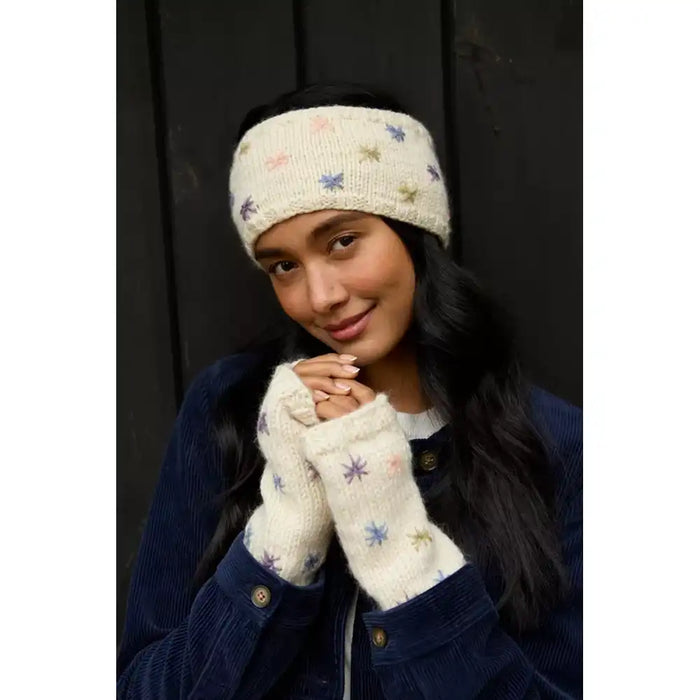 A woman wearing a cream-coloured knit headband and matching fingerless gloves with small multicolored star patterns, paired with a dark blue corduroy jacket against a dark wooden background.