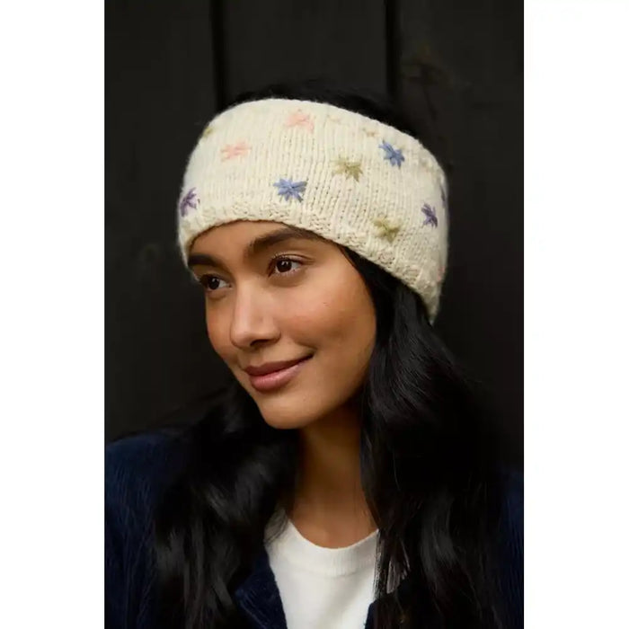 A woman wearing a cream-colored knit headband with small pastel embroidered flowers in blue, pink, and green, set against a dark background.