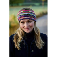 A woman wearing a colorful striped knit headband with a small brown leather brand patch, photographed outdoors on a blurred path.