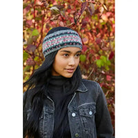A woman wearing a headwarmer with a multicolor Fair Isle pattern of pink, blue, and grey, styled with a black turtleneck and a dark denim jacket against a background of autumn foliage.