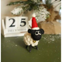 A small handmade felt sheep decoration wearing a red Santa hat and gold bell, standing on a dark green surface with a blurred calendar and holiday decor in the background.