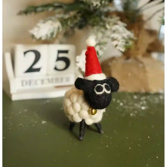 A small handmade felt sheep decoration wearing a red Santa hat and gold bell, standing on a dark green surface with a blurred calendar and holiday decor in the background.