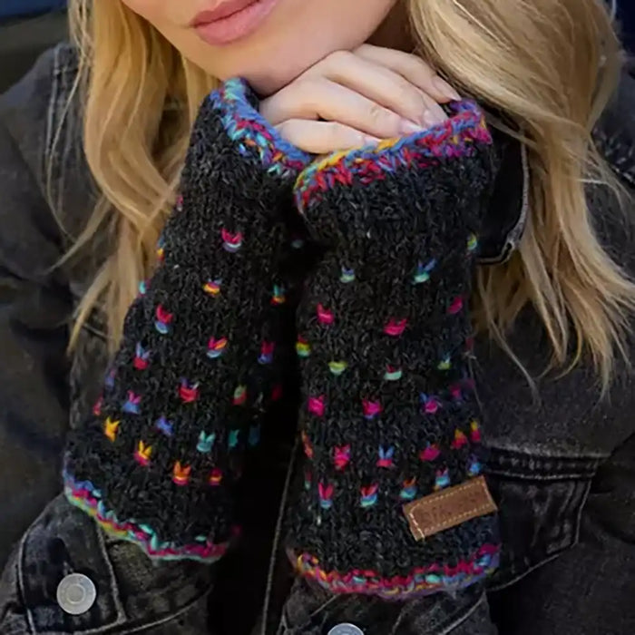 Close-up of a person wearing black knit fingerless wrist warmers with multicolored yarn accents and a small brown leather patch, paired with a dark denim jacket.