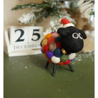 A handmade felt sheep figurine with a black wool body, a colorful pom-pom wool fleece, a red Santa hat, and a small gold bell, standing on a dark green surface with a wooden calendar in the background.
