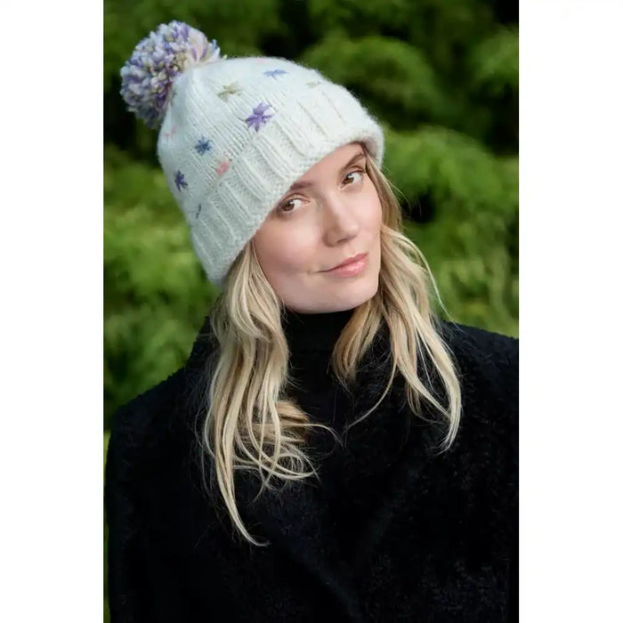 A woman wearing a cream-coloured knit beanie with a pom-pom and small purple and pink star patterns, set against a blurred green background.