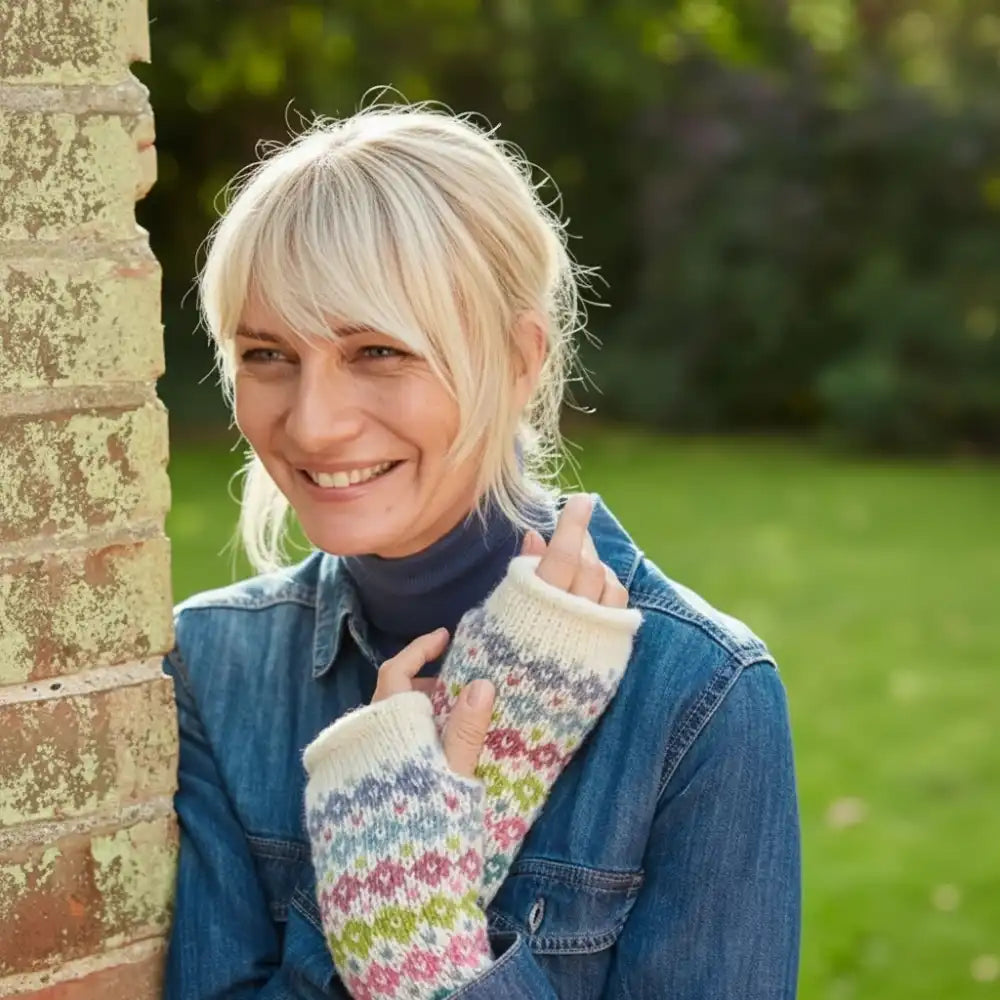 A smiling blonde woman outdoors leaning against a brick pillar, wearing a blue denim jacket over a dark navy turtleneck and cream knit fingerless gloves with a multicolor fair isle pattern.