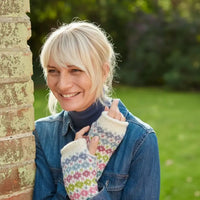 A smiling blonde woman outdoors leaning against a brick pillar, wearing a blue denim jacket over a dark navy turtleneck and cream knit fingerless gloves with a multicolor fair isle pattern.
