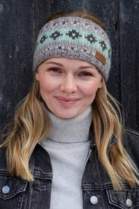 A woman wearing a grey knit headband with a mint green and black Fair Isle pattern, styled with a grey turtleneck sweater and a dark denim jacket against a dark wooden background.