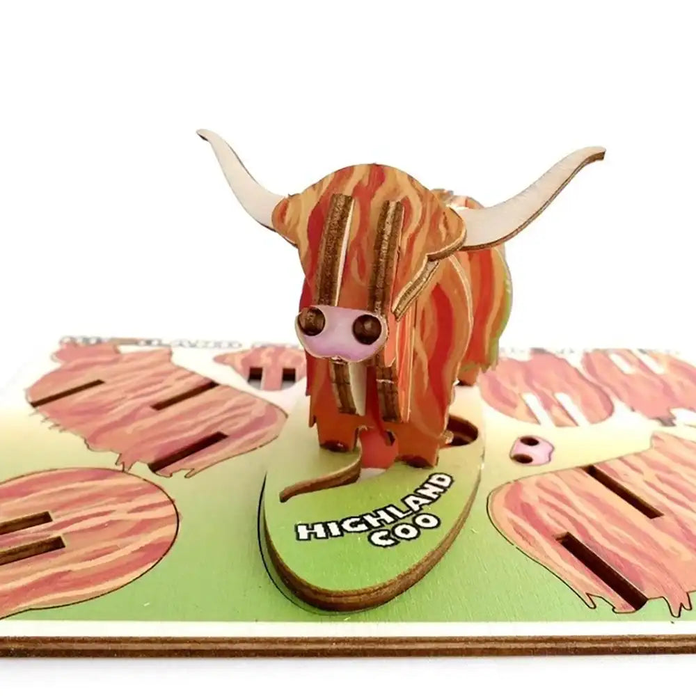 A 3D wooden pop-up model of a Highland Coo, featuring a striped orange and brown body with cream horns, standing on a green base labeled 'HIGHLAND COO' with additional cutout landscape details.