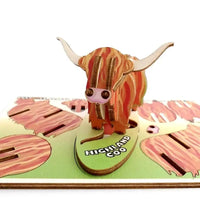 A 3D wooden pop-up model of a Highland Coo, featuring a striped orange and brown body with cream horns, standing on a green base labeled 'HIGHLAND COO' with additional cutout landscape details.