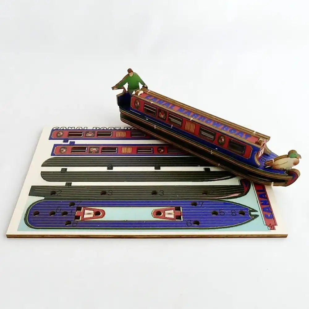 A wooden 3D puzzle model of a canal boat, shown assembled and resting diagonally on top of its flat printed wooden base, photographed against a clean white background.