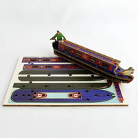 A wooden 3D puzzle model of a canal boat, shown assembled and resting diagonally on top of its flat printed wooden base, photographed against a clean white background.