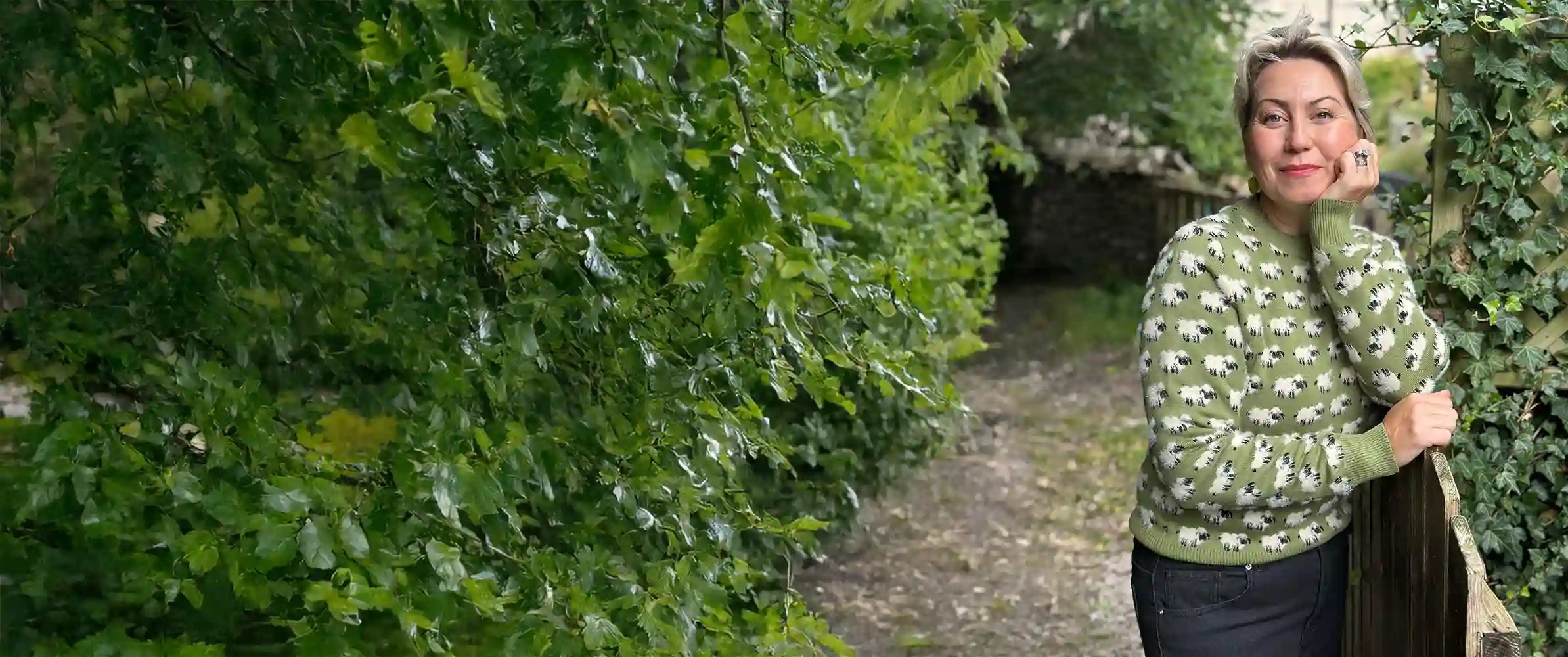 A woman wearing a green and white patterned knit sweater and dark trousers stands on a garden path surrounded by dense green foliage and ivy.