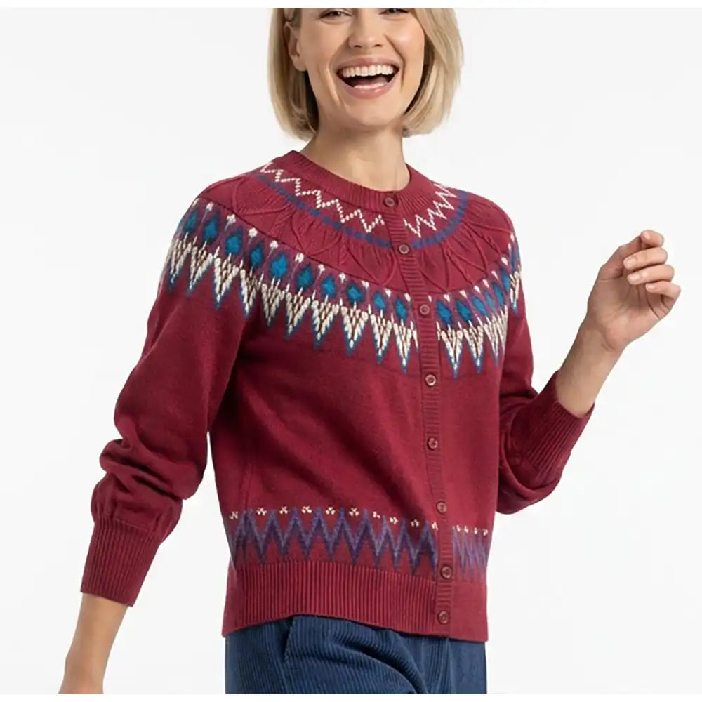 A woman wearing a burgundy button-up cardigan with a colorful geometric Fair Isle pattern across the chest and hem, paired with blue corduroy pants against a white studio background.