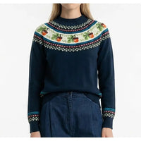 A woman wearing a navy blue knit sweater with a colorful Fair Isle style yoke pattern featuring green leaves, red berries, and white geometric borders, paired with dark blue jeans against a plain white background.
