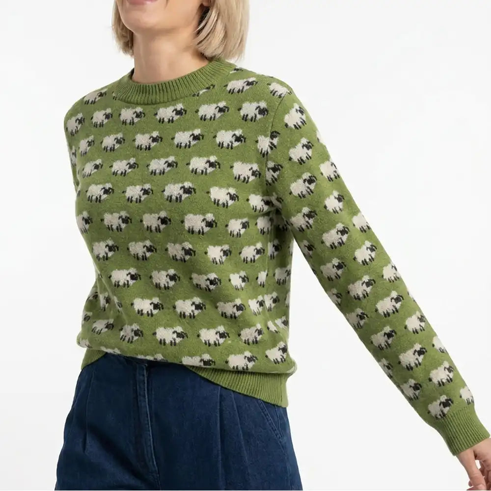 A woman wearing a sage green crewneck sweater with an all-over repeating pattern of black-and-white sheep, paired with dark blue jeans against a plain white background.