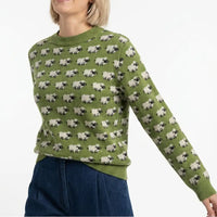 A woman wearing a sage green crewneck sweater with an all-over repeating pattern of black-and-white sheep, paired with dark blue jeans against a plain white background.