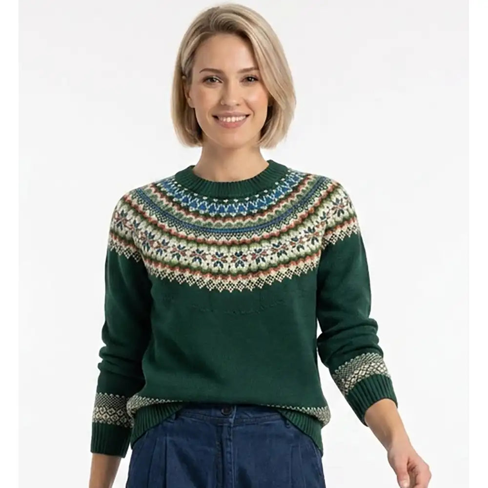 A woman wearing a dark green crewneck sweater with a colorful Fair Isle pattern across the yoke and cuffs, paired with blue jeans, against a plain white background.