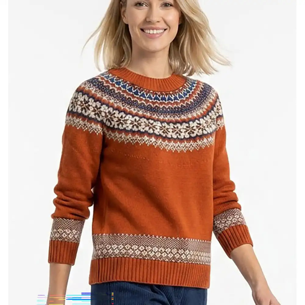 A woman wearing a rust-orange Fair Isle knit sweater with a multicolor patterned yoke and cuffs, paired with dark blue jeans, against a plain white background.