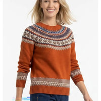 A woman wearing a rust-orange Fair Isle knit sweater with a multicolor patterned yoke and cuffs, paired with dark blue jeans, against a plain white background.