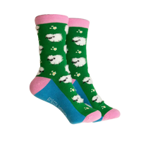 A pair of crew socks with a green background, white sheep pattern, pink cuffs and heels, and a blue toe section. The brand name 'pretty socks' is printed on the side of the blue toe area.