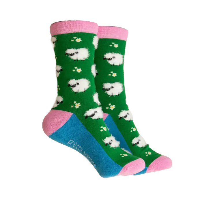 A pair of crew socks with a green background, white sheep pattern, pink cuffs and heels, and a blue toe section. The brand name 'pretty socks' is printed on the side of the blue toe area.