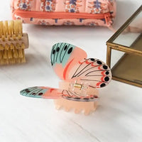 Butterfly-shaped hair clip on a white surface with a small bag and brush in the background.