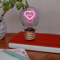 Light bulb with heart-shaped neon light on a red book, surrounded by a plant and stationery items.