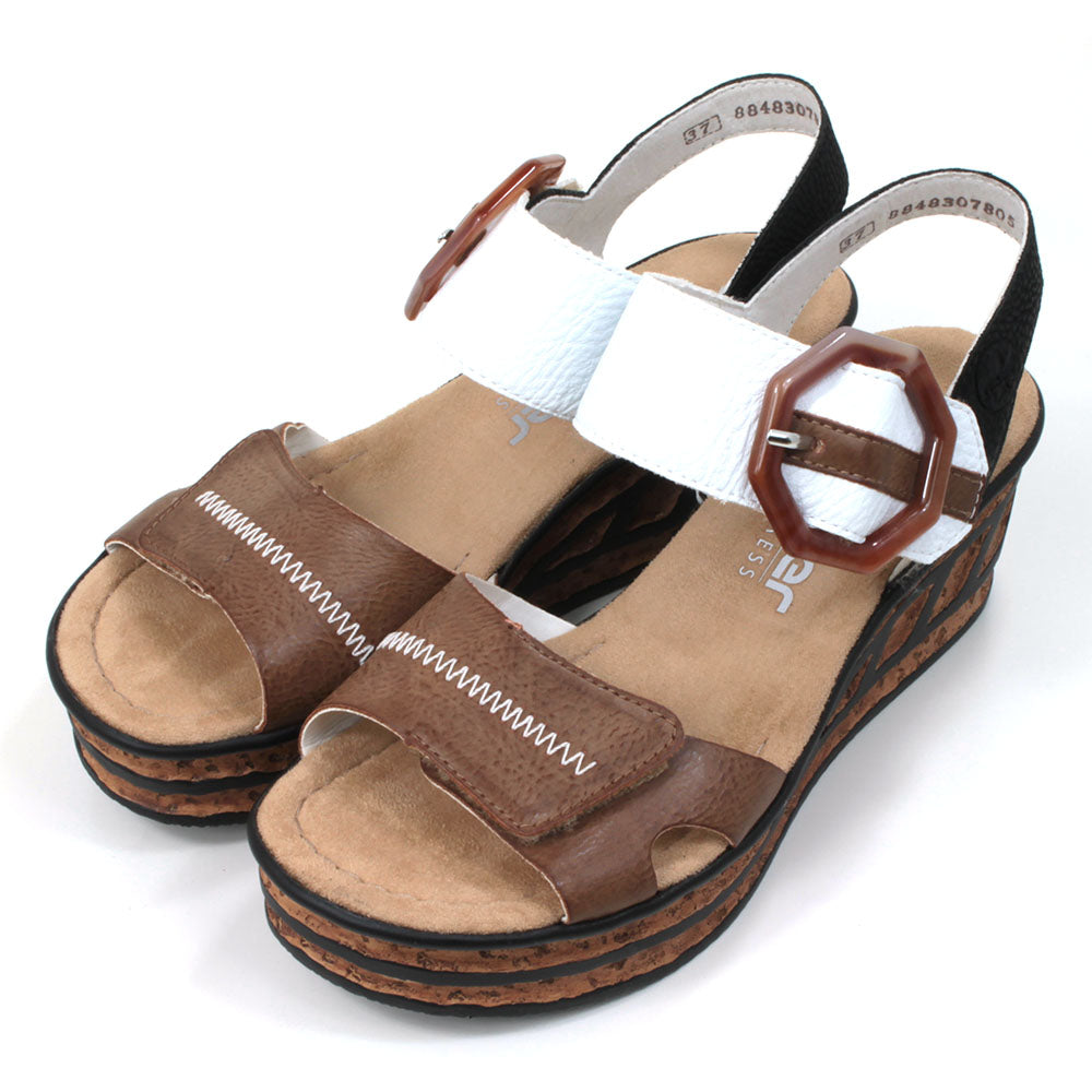 Rieker cork wedge sandals. Velcro fitting and adjustment. Brown over toes strap. White over upper foot strap with brown buckle detail. Black strap around the ankle. Tan insoles. Angled view.