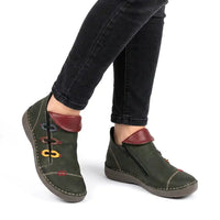 A pair of dark olive green ankle boots with reddish-brown faux leather collar accents and colorful embroidered details, shown on a model wearing black jeans against a white background.