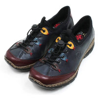 Rieker dark blue leisure shoes. Black fabric padding around the ankle and over the top of the tongue. Blue, yellow and brown details around the first,third and fifth lace eyes. Burgundy strips around the toes.  Fastens with elastic laces which are adjusted with a push button gripper. Cork and black rubber soles. Angled view.