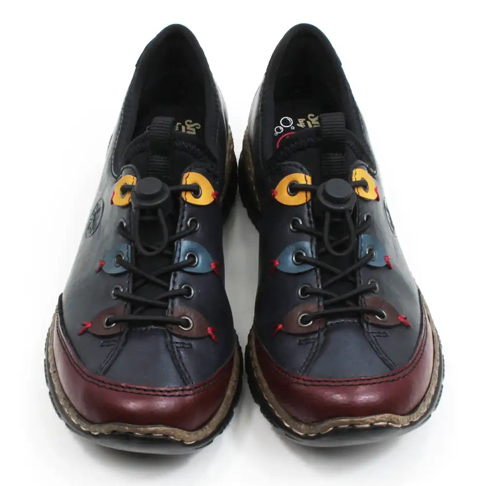 Rieker dark blue leisure shoes. Black fabric padding around the ankle and over the top of the tongue. Blue, yellow and brown details around the first,third and fifth lace eyes. Burgundy strips around the toes.  Fastens with elastic laces which are adjusted with a push button gripper. Cork and black rubber soles. Front view.