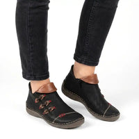 A pair of black faux leather ankle boots with brown faux leather trim and decorative brown-and-red embroidered details, shown on a model wearing black jeans against a white background.