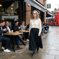 A woman wearing a cream button-up shirt and a long black skirt walks along a city sidewalk in front of a coffee shop named The Daily Grind. Patrons are seated at outdoor tables, and a red double-decker bus is visible in the background.
