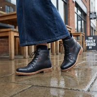 A person wearing black leather lace-up ankle boots with tan soles, standing on wet pavement outdoors.