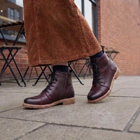 A person wearing dark burgundy lace-up ankle boots with a tan sole, paired with brown corduroy trousers, standing on a stone patio in front of a brick building.