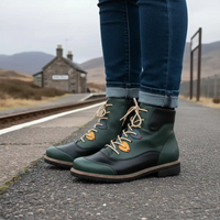 A person wearing dark green and black lace-up ankle boots with tan laces and yellow accents, standing on a train station platform with cuffed blue jeans and a blurred station building in the background.