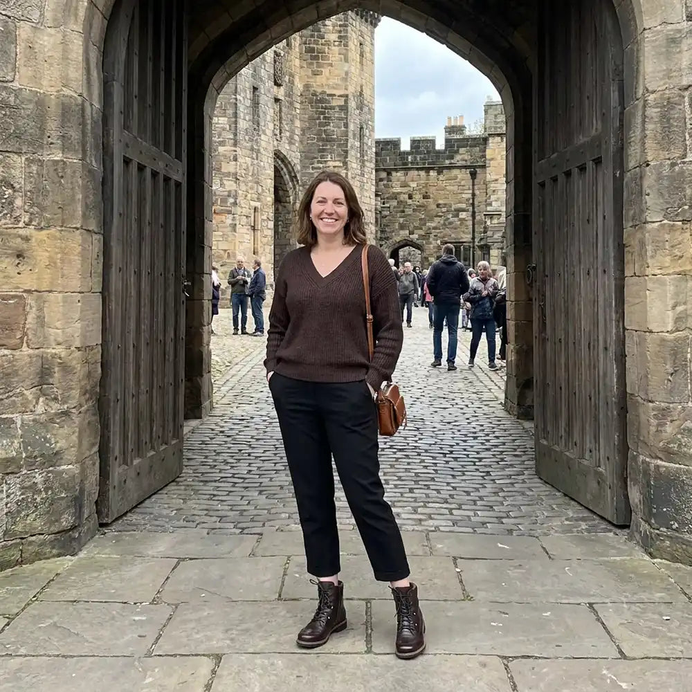 A woman stands in front of a large stone archway in an old castle courtyard, wearing a brown V-neck sweater, black cropped pants, brown ankle boots, and carrying a small brown shoulder bag.
