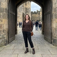 A woman stands in front of a large stone archway in an old castle courtyard, wearing a brown V-neck sweater, black cropped pants, brown ankle boots, and carrying a small brown shoulder bag.