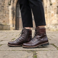 A pair of dark brown leather lace-up ankle boots with side zippers, worn with black trousers on a stone pavement in front of a historic stone building.