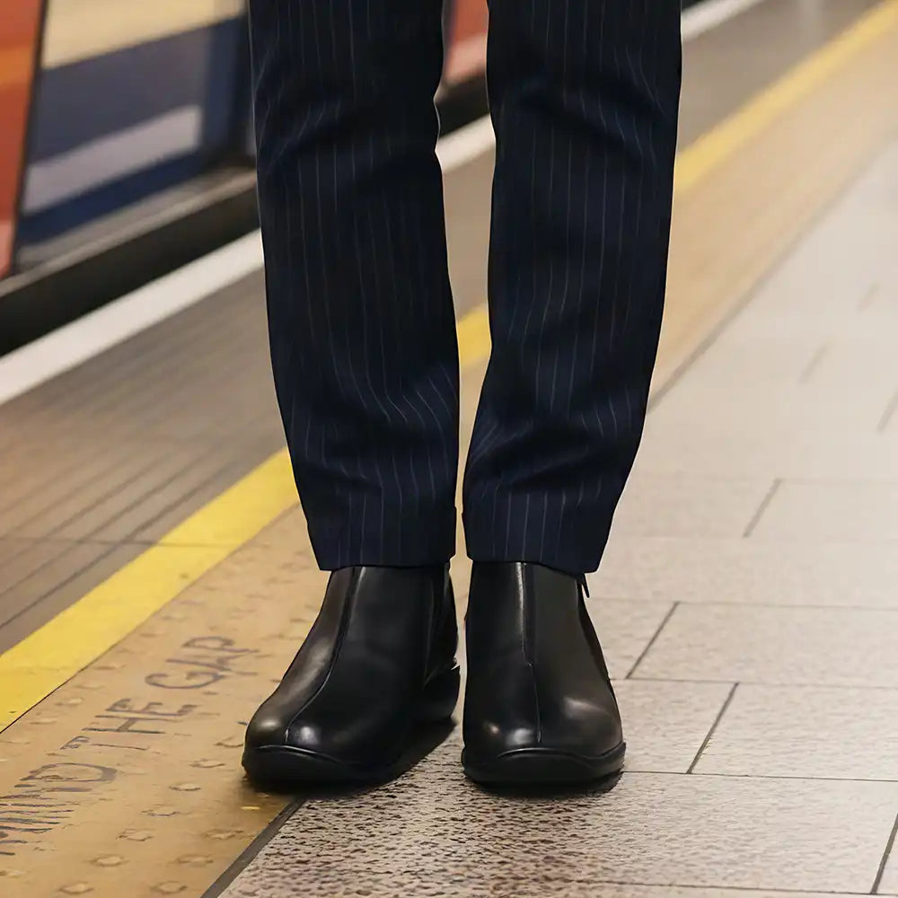 A close-up of a person's legs wearing dark navy pinstripe trousers and black Chelsea boots on a subway platform.