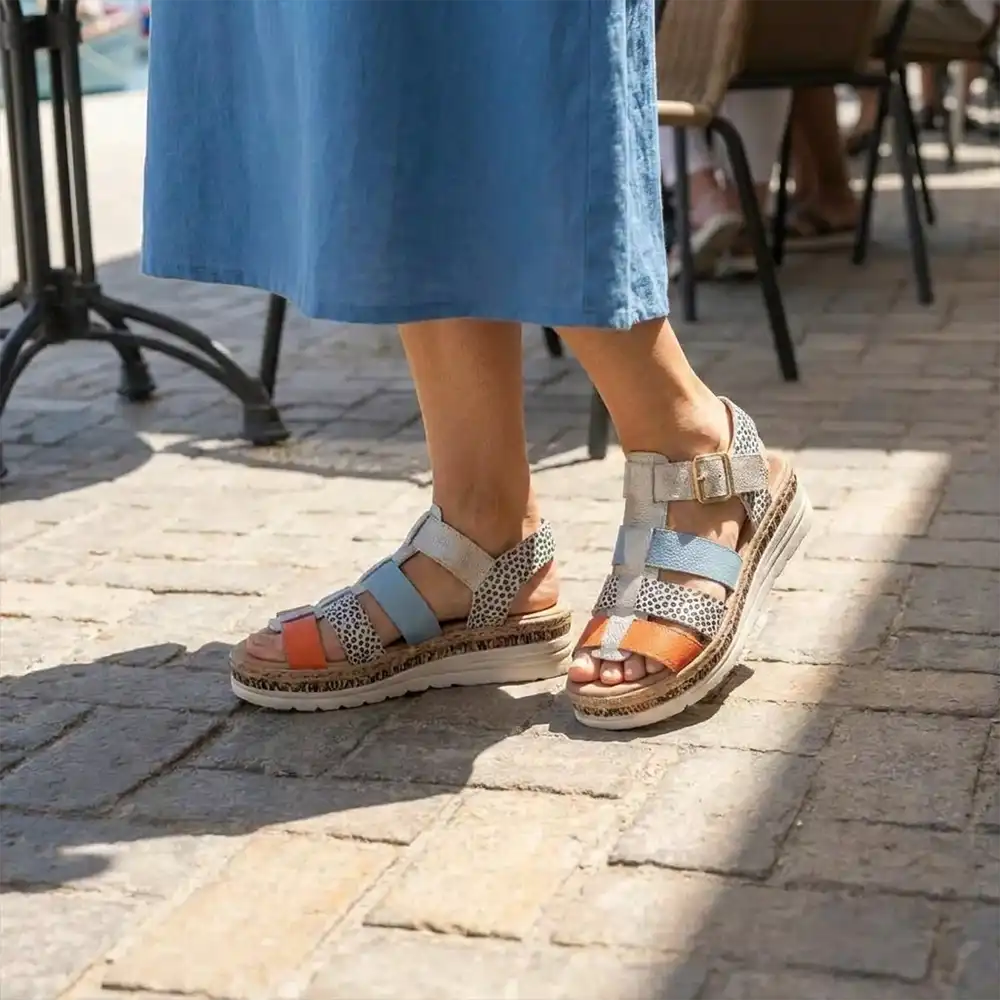 A person wearing light blue wedge sandals with patterned straps and orange accents, standing on a sunlit stone pavement near outdoor cafe chairs.