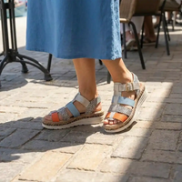 A person wearing light blue wedge sandals with patterned straps and orange accents, standing on a sunlit stone pavement near outdoor cafe chairs.