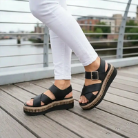 Black platform wedge sandals with wide crisscross straps and a buckle ankle strap, shown on a person wearing white pants standing on a wooden deck outdoors.