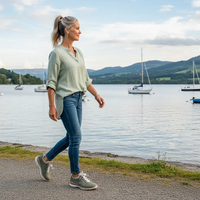 Woman in green blouse, blue jeans and green trainers walking beside a lake.